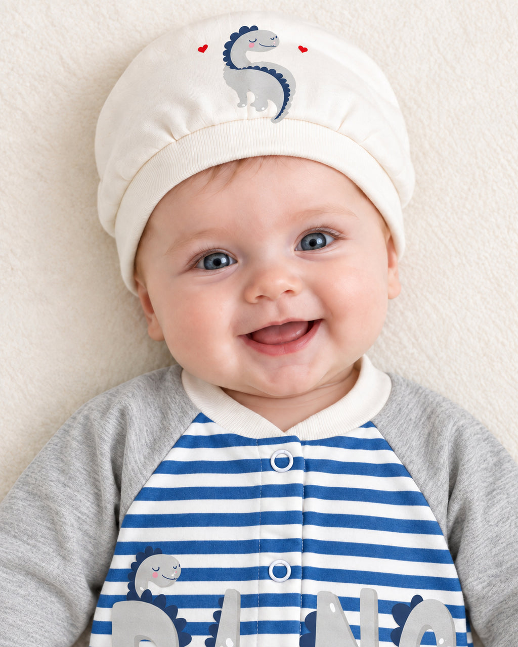 Baby Dream grey & blue dinosaur sleepsuit and hat set, close baby portrait with matching dinosaur cap
